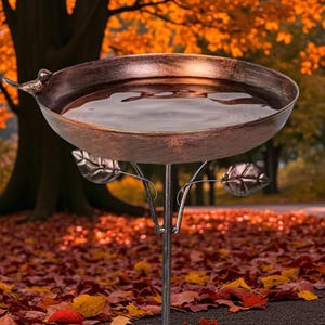 May include: A bronze-effect bird bath with a bird detail on the rim. The bowl is filled with water and supported by a metal stand with leaf accents. The background shows autumn leaves.