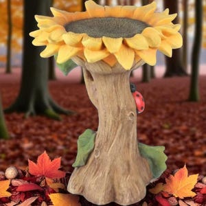 Puede incluir: Un comedero decorativo para pájaros con forma de girasol. Los pétalos amarillos rodean un centro gris oscuro. El comedero está sostenido por un tronco de árbol con una mariquita y hojas verdes. El comedero para pájaros está hecho de un material duradero.
