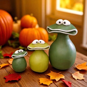 May include: Three whimsical green frog figurines with white-rimmed eyes and mushroom-shaped heads. The frogs are displayed on a wooden surface with colorful autumn leaves and pumpkins in the background. The figurines vary in size.