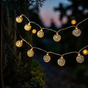 May include: A string of decorative globe lights featuring spherical bulbs with a metallic, perforated finish. Each sphere emits a warm, golden glow through its intricate cut-out pattern, connected by a thin, dark wire arranged in two distinct loops.