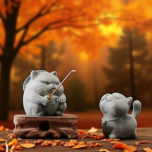 Könnte beinhalten: Zwei graue Katzenfiguren. Eine Katze sitzt auf einem Holzblock und hält einen Stock. Die andere Katze steht mit geschlossenen Augen. Der Hintergrund zeigt eine unscharfe Herbstszene mit orangefarbenen Blättern.