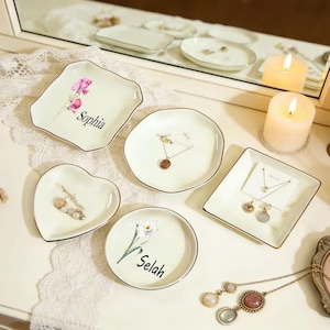 Puede incluir: Una colección de platos de cerámica blanquecina para joyas en varias formas, incluyendo cuadrada, corazón y redonda. Cada plato tiene un borde dorado y presenta un diseño floral y un nombre. Se exhiben joyas en algunos de los platos.