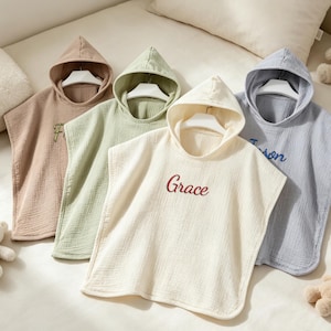May include: Four baby ponchos with hoods in muted tones: beige, green, cream, and blue. Each poncho features a hood and an embroidered name in a contrasting colour. The cream poncho is embroidered with "Grace" in red.