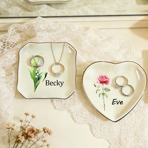 Puede incluir: Dos platos decorativos para anillos. Uno es cuadrado con un diseño floral y el nombre "Becky". El otro tiene forma de corazón con una rosa y el nombre "Eve". Ambos platos tienen ribete dorado y sostienen anillos y un collar.