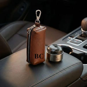 May include: A brown leather key pouch with a silver zip and clasp, with the initials "BC". Beside it is a silver and black car air freshener. The items are on a brown leather car armrest. The car interior is visible in the background.