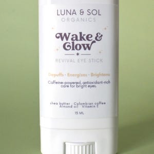 Op de afbeelding: Een witte, rechthoekige oogstick met de tekst "LUNA & SOL ORGANICS Wake & Glow REVIVAL EYE STICK". De stick bevat de tekst "Depuffs. Energizes. Brightens" en "Caffeine-powered, antioxidant-rich care for bright eyes." Het bevat 15 ml.