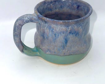 May include: A handmade ceramic mug with a unique shape and color scheme. The mug features a blue and gray speckled glaze on the upper portion, transitioning to a teal green base. The handle is curved and matches the upper glaze.