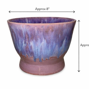 May include: A handcrafted ceramic planter with a wide, flared rim and a pedestal base. The planter is approximately 8 inches wide and 6 inches tall. It features a blend of purple, blue, and brown glaze, creating a unique, artistic appearance.