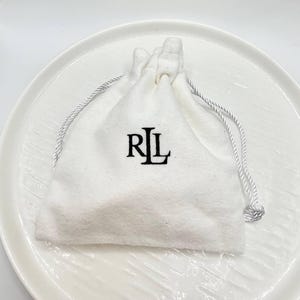 May include: A white drawstring pouch with the letters "RL" in black, resting on a white, round plate. The pouch has a silver-colored drawstring closure. The background is a plain white surface.