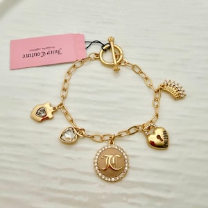 Könnte beinhalten: Goldfarbenes Charm-Armband mit verschiedenen Anhängern, darunter ein Herz, eine Krone und ein runder Anhänger mit den Initialen "JC". Das Armband hat einen Knebelverschluss und ein rosa Etikett mit "Juicy Couture".