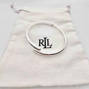 May include: A silver-coloured, open cuff bracelet with a smooth, polished finish. The bracelet is displayed on a white drawstring pouch with the letters "RL" in black. The bracelet has a curved design.