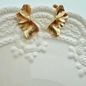 May include: A pair of gold-coloured stud earrings. Each earring has a detailed, textured design resembling a stylized wing or leaf. The earrings are displayed on a white, patterned surface.