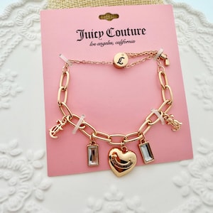 Könnte beinhalten: Ein goldfarbenes Juicy Couture Charm-Armband auf einer rosa Karte. Das Armband hat ein Kettenglied-Design mit rechteckigen und herzförmigen Charms. Auf der Karte stehen der Markenname und der Standort.