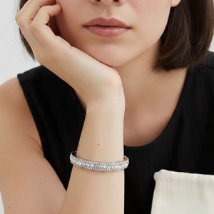 Puede incluir: Un brazalete plateado con múltiples filas de cristales transparentes y brillantes. El brazalete es de estilo brazalete y los cristales están muy juntos. El brazalete se lleva en una muñeca, con un top negro de fondo.
