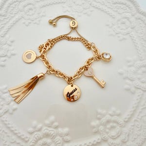 May include: Gold-toned charm bracelet with multiple charms, including a heart, a key, a tassel, and a round charm with the word "Guess". The bracelet has a sliding clasp for adjustable sizing.