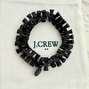 Op de afbeelding: Zwart kralenarmband met een uniek ontwerp, op een witte stoffen tas. De tas heeft de tekst "J.CREW" in het groen. De armband is gemaakt van kleine, kegelvormige kralen.