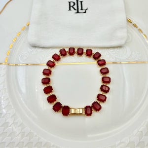 May include: A gold bracelet featuring rectangular, deep red gemstones. The bracelet is presented on a white plate with a gold rim, alongside a white pouch with the letters "RL" in black. The bracelet is secured with a gold clasp.