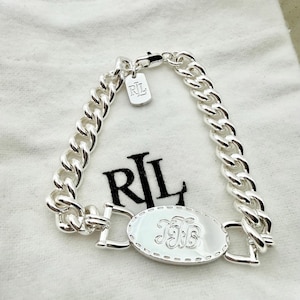 Könnte beinhalten: Ein silberfarbenes Armband mit einem Panzerketten-Design. Das Armband hat ein ovales Mittelstück mit einem eingravierten Monogramm und ein kleines Etikett mit den Buchstaben "RL". Das Armband wird auf einem weißen Stoffbeutel präsentiert.