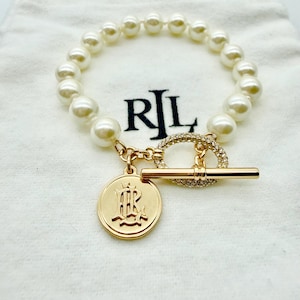 May include: A pearl bracelet with a gold-tone toggle clasp and a circular charm. The charm features an embossed design and the letters "RL". The bracelet is displayed on a white drawstring bag with the letters "RL" printed on it.
