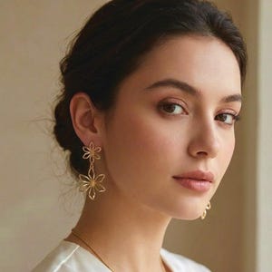 May include: Gold-coloured flower earrings. The earrings have a delicate, openwork design with two flower shapes, one above the other. The earrings are dangling and are worn by a person with dark hair and a white top.