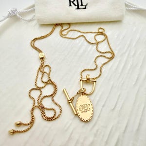 May include: Gold-coloured necklace with an oval pendant featuring engraved initials. The necklace has a delicate chain and a toggle clasp. A small white pouch with the letters "RL" in black is in the background.