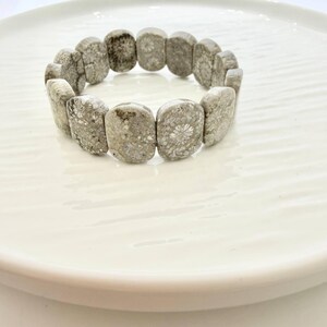Puede incluir: Un brazalete de coral fosilizado gris y blanco. El brazalete está hecho de piedras rectangulares pulidas con patrones naturales. El brazalete se exhibe en un plato blanco.