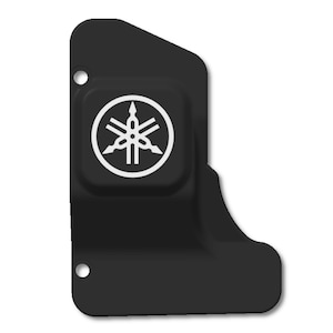May include: A black, irregularly shaped component with a white Yamaha logo. The logo is a stylized emblem within a square, centered on the component. Two small holes are visible. The component has a smooth, matte finish.