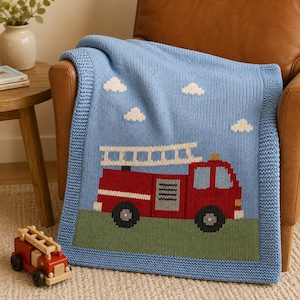 May include: A light blue knitted blanket featuring a red fire truck with a white ladder on top, driving on a green field under a sky with white clouds. The blanket has a textured border and is displayed alongside a small wooden toy fire truck.