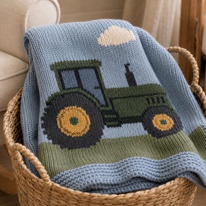 Puede incluir: Una manta de punto azul claro con un tractor verde de ruedas amarillas y una nube blanca. La manta tiene un patrón de tejido texturizado y está doblada dentro de una cesta tejida. El diseño incluye un campo verde en el borde inferior.