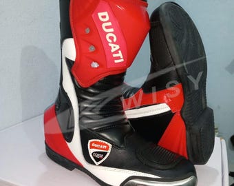 May include: Motorcycle boots in black, red, and white, featuring the "Ducati" logo. These high-top boots have a protective shin guard and a reinforced toe. They include a metal toe plate and are designed for motorcycle use.