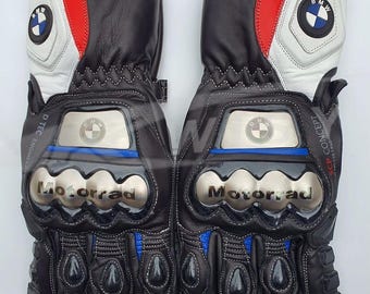 Może przedstawiać: Para czarnych skórzanych rękawic motocyklowych z białymi, czerwonymi i niebieskimi akcentami. Posiadają logo BMW i napis "Motorrad" w kolorze srebrnym. Zawierają ochraniacze kostek i palców.