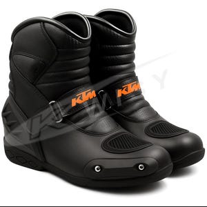 Puede incluir: Par de botas de moto negras con logotipos KTM naranjas. Las botas son de media caña, con cierre de correa y paneles reforzados. Las suelas son negras y tienen un diseño texturizado.