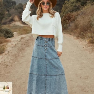 Könnte beinhalten: Eine Frau trägt einen weißen, gecroppten Pullover, eine Sonnenbrille und einen braunen Hut. Sie trägt einen langen, hellblauen Jeansrock mit Knöpfen. Das Outfit ist lässig und für den Outdoor-Bereich geeignet.