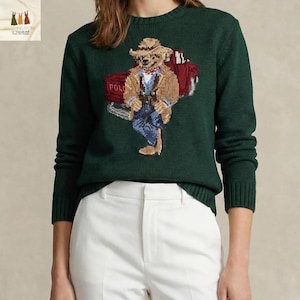 Puede incluir: Un suéter verde oscuro con un diseño de oso vaquero. El oso lleva sombrero, chaqueta y vaqueros, y está de pie frente a un carro rojo. La palabra "POLO" es visible en el carro.