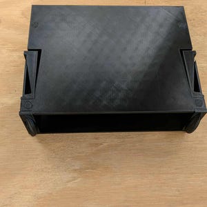 May include: A black, rectangular, 3D-printed object with a textured surface. The object has a box-like shape with rounded corners and small wheels on the sides. It sits on a light brown wooden surface.