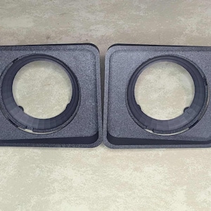 May include: Two dark gray, square-shaped speaker enclosures with circular openings. The enclosures have a textured surface and are designed to house speakers. The openings are surrounded by a raised, rounded edge.