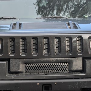 May include: A black Hummer vehicle front grille featuring seven vertical slots with a honeycomb mesh pattern. The word HUMMER is embossed across the center of the grille. Below the main grille is a secondary horizontal intake with a circular mesh.
