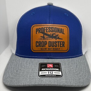 May include: A blue, white, and gray trucker hat with a brown leather patch. The patch reads "PROFESSIONAL CROP DUSTER" with an image of a crop duster plane. The hat has a gray brim and a white mesh back. The hat is adjustable.