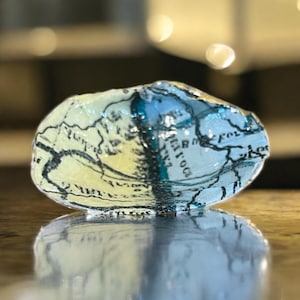 May include: A decorative seashell, hand-painted with a map design. The shell displays light blue, yellow, and black hues. The map design is visible across the surface, adding a unique artistic touch to the seashell.