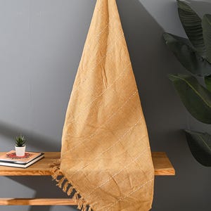 May include: Mustard yellow throw blanket with white stitched lines and fringe detail. The blanket is draped over a wooden bench, with a small potted plant and a book visible. The background is a grey wall.
