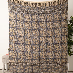 May include: A large, rectangular blue and beige floral patterned throw blanket with fringe trim. The blanket features a repeating floral design and a decorative border. The top and bottom edges have a natural-coloured fringe.