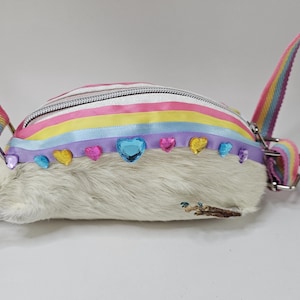 Può includere: Una borsa a tracolla a forma di ratto, con un corpo in finta pelliccia bianca e una parte superiore a righe arcobaleno. La borsa è adornata con abbellimenti a forma di cuore e gemme. Ha una tracolla arcobaleno e una coda glitterata.