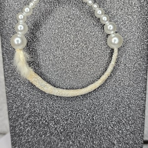 May include: A pearl bracelet with a toggle clasp. The bracelet features a series of small and large white pearls. The curved portion of the bracelet is covered in white glitter and has a small white feather attached.