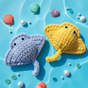 Könnte beinhalten: Zwei gehäkelte Rochen, einer hellblau und einer gelb, mit schwarzen Knopfaugen, befinden sich auf einem blauen Hintergrund mit Muscheln und Perlen. Die Rochen sind der Mittelpunkt, umgeben von dekorativen Elementen.