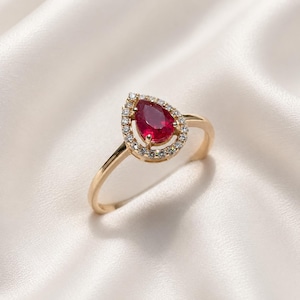 May include: A gold ring featuring a teardrop-shaped, deep red gemstone, encircled by a halo of small, clear gemstones. The ring's band is slender and gold. The ring is displayed on a white, silky backdrop.