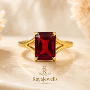 Puede incluir: Un anillo de oro con una gran piedra preciosa rectangular de color rojo intenso en un engaste de cuatro garras. La banda tiene un diseño de vástago dividido que se estrecha hacia la piedra central. El metal tiene un acabado pulido y facetado.