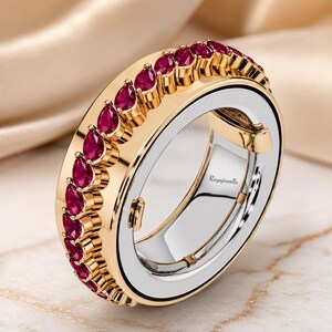 Puede incluir: Un anillo de oro con una hilera de rubíes rojos en talla pera a lo largo del borde exterior. La banda interior es de tono plateado con el texto Rayajewells grabado en la superficie. El diseño incluye un mecanismo de bisagra en el lateral.