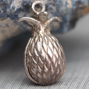 May include: A small, silver-colored pineapple charm with detailed texture. The charm has a loop at the top for attachment and is likely made of metal. The pineapple design is a classic symbol of hospitality.