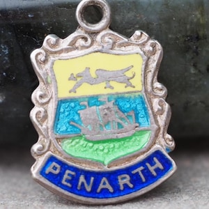 May include: A silver-toned Penarth charm with a shield design. The shield features a yellow top with a camel, a blue center with a ship, and a green base. The word "PENARTH" is in blue on a curved banner.