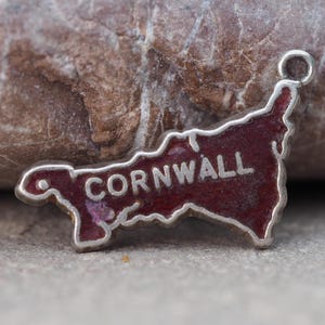 May include: A silver-toned charm in the shape of Cornwall, with a dark red enamel fill. The word "CORNWALL" is inscribed in silver across the center. The charm has a small loop at the top for attachment.